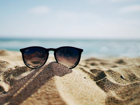 Black Ray-Ban sunglasses resting on a mound of sand at a sunny beach with the sea blurred in the background.